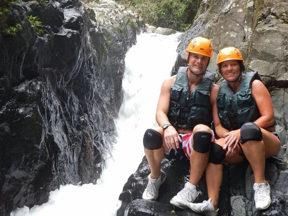 Couple in helmets and life jackets seated on the rock beside a waterfall pool, El Yunque